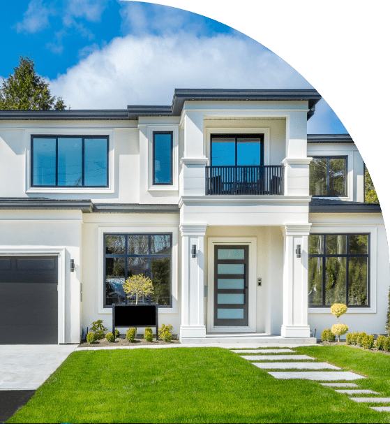 Modern two-story house with large windows, white exterior, and a dark garage door. Neatly trimmed grass and shrubs line the front yard under a partly cloudy sky.