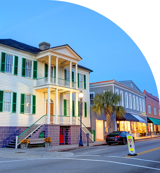 A street view of historic buildings at dusk, featuring a large two-story house with green shutters and a shopfront. A pedestrian crossing sign is visible in the foreground.