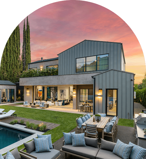Modern house with a large patio, outdoor seating, and a pool. The sky shows a vibrant sunset.