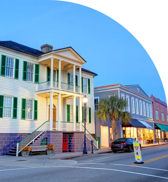 A street view of historic buildings at dusk, featuring a large two-story house with green shutters and a shopfront. A pedestrian crossing sign is visible in the foreground.